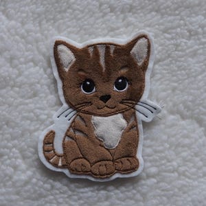 Könnte beinhalten: Eine gestickte Katzen-Applikation in Braun und Weiß auf weißem Hintergrund. Die Katze sitzt mit den Pfoten unter dem Körper und hat einen weißen Fleck auf der Brust.