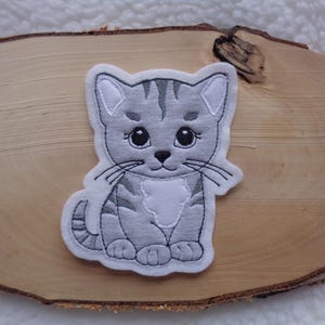 Könnte beinhalten: Ein grau-weißer Katzenaufnäher mit einem niedlichen Design. Die Katze hat große schwarze Augen, Schnurrhaare und gestreifte Markierungen. Der Aufnäher befindet sich auf einer Holzoberfläche mit hellem Hintergrund.