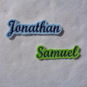Könnte beinhalten: Zwei gestickte Stoffpatches mit Namen. Der obere Patch ist hellblau mit dem Namen "Jonathan" in dunkelblauem Garn. Der untere Patch ist grün mit dem Namen "Samuel" in dunkelblauem Garn.