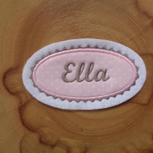 Könnte beinhalten: Ein weißer ovaler Stoff-Patch mit braunem gewelltem Rand. Der Patch hat einen rosa Oval im Inneren mit weißen Punkten und den Namen "Ella" in Braun gestickt.