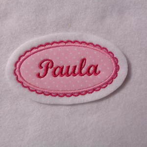 Könnte beinhalten: Ein rosa ovaler Stoff-Patch mit weißem Punktmuster und dem Namen "Paula" in rosa gestickt.