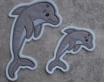 Könnte beinhalten: Zwei graue gestickte Delfin-Patches mit weißen Umrandungen. Der größere Delfin springt aus dem Wasser und der kleinere Delfin schwimmt.