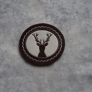 Könnte beinhalten: Ein braunes gesticktes Patch mit weißem Hintergrund, das eine Silhouette eines Hirsches mit Geweih zeigt. Das Patch ist rund und hat einen dekorativen Rand.