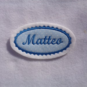 Könnte beinhalten: Weißer ovaler Stoff-Patch mit blauem gewelltem Rand und weißen Punkten. Der Name "Matteo" ist in blauer Kursivschrift gestickt.