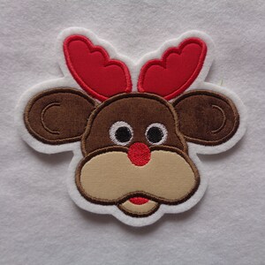 Könnte beinhalten: Gestickter Rentier-Gesicht-Aufnäher mit braunen, roten, beigen und weißen Details. Das Rentier hat große braune Ohren, rote Geweihe, eine beige Schnauze und eine rote Nase. Der Aufnäher ist weiß umrandet.