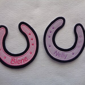Könnte beinhalten: Zwei gestickte Hufeisen-Patches, einer rosa mit dem Namen "Biene" und einer lila mit dem Namen "Nelly".