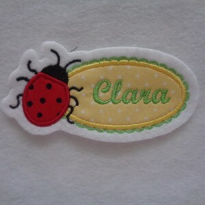 Könnte beinhalten: Ein weißer Stoffoval mit grünem gewelltem Rand und gelber Mitte mit weißen Punkten. Der Name "Clara" ist in Grün in den Oval gestickt. Eine rote Marienkäfer mit schwarzen Punkten ist links neben dem Oval gestickt.
