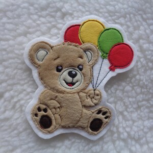 Könnte beinhalten: Ein beigefarbener Plüsch-Teddybär-Aufnäher mit dunkelbraunen Tatzen und einer schwarz gestickten Nase. Der Bär hält ein Bündel aus vier Filzballons in Rot, Gelb und Grün, die mit dünnen schwarzen Fäden befestigt sind, auf weißem Filzhintergrund.