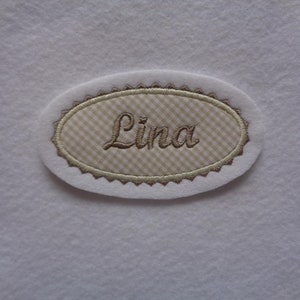 Könnte beinhalten: Ein weißer ovaler Stoff-Patch mit einem braunen gestickten Rand und einem Gingham-Muster im Inneren. Der Name "Lina" ist in brauner Kursivschrift gestickt.