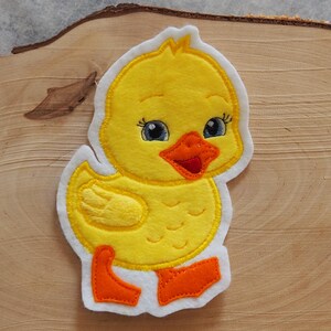 Könnte beinhalten: Ein gelbes Entlein-Applikation mit weißem Rand. Das Entlein hat blaue Augen, einen orangefarbenen Schnabel und Füße sowie einen gelben Körper. Die Applikation ist aus Filz und dient wahrscheinlich zum Basteln oder Verzieren.