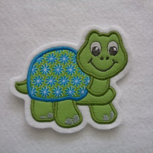 Könnte beinhalten: Grünfilz-Applikation einer lächelnden Schildkröte mit blau-grünem Blumenmuster auf ihrem Panzer.