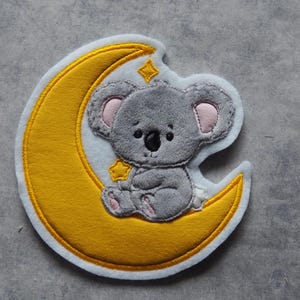 Könnte beinhalten: Ein grauer Koalabär sitzt auf einem gelben Halbmond mit weißen Nähten. Der Mond hat einen weißen Hintergrund und gelbe Sterne.