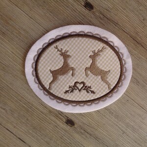 Könnte beinhalten: Ein weißer ovaler Untersetzer mit einem braunen gestickten Design aus zwei Rehen mit einem Herz und Blättern in der Mitte. Der Untersetzer hat ein Gingham-Muster und einen gewellten Rand.