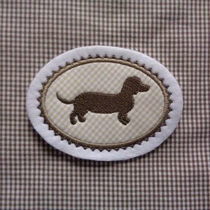 Könnte beinhalten: Ein brauner Dackel-Silhouetten-Stick auf einem beige-weißen Gingham-Stoff-Patch. Der Patch ist oval geformt und hat einen weißen Rand mit einem braunen gewellten Rand.