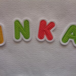 Könnte beinhalten: Ein Filznamenbanner mit dem Namen "INKA" in bunten Buchstaben. Die Buchstaben sind grün, gelb, rosa und grün mit weißen Punkten.
