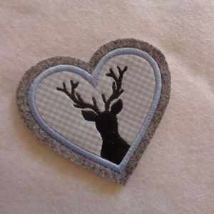 Könnte beinhalten: Ein grauer Filz-Herz-Patch mit einem blauen und weißen Gingham-Muster im Inneren. Eine schwarze Silhouette eines Hirsches mit Geweih ist auf das Gingham gestickt.