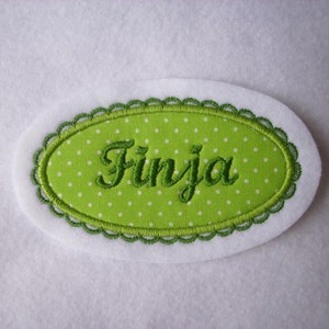 Könnte beinhalten: Grüner ovaler Stoff-Patch mit weißen Punkten und dem Namen "Finja" in grüner Stickerei.
