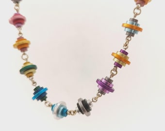 May include: A colorful necklace featuring a series of stacked, disc-shaped beads in various colors, including purple, pink, orange, and blue. The beads are connected by a silver chain, creating a vibrant and eye-catching accessory.