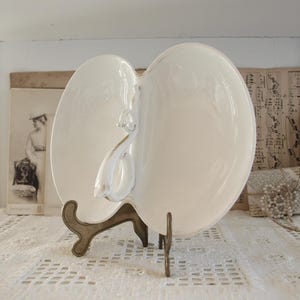 May include: An off-white, heart-shaped serving dish with a swan handle, displayed on a decorative metal stand. The background features antique ephemera and a lace tablecloth. The dish's design and presentation suggest a vintage, elegant style.