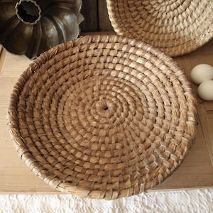 Op de afbeelding: Een grote, ronde, geweven mand gemaakt van natuurlijke materialen. De mand is bruin en heeft een cirkelvormig patroon. Hij staat op een houten oppervlak.