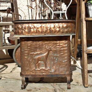 Può includere: Un vaso di rame con un disegno di cervo e un bordo floreale. Il vaso ha un aspetto rustico ed è fatto di metallo.