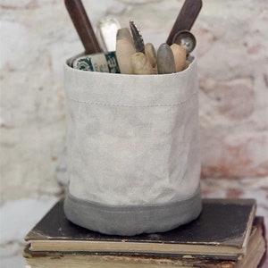 Könnte beinhalten: Ein weißer Canvas-Aufbewahrungskorb mit grauem Besatz, gefüllt mit verschiedenen Werkzeugen und Utensilien, der auf einem Stapel alter Bücher steht.