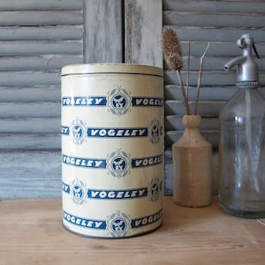 Könnte beinhalten: Eine vintage Blechdose mit einem blau-weißen Muster, auf dem der Text "VOGEL EY" wiederholt um die Dose herumläuft. Die Dose steht auf einer Holzoberfläche.