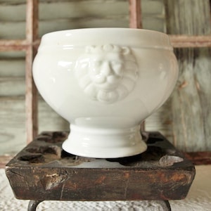 Puede incluir: Un cuenco de cerámica blanca con un emblema de cabeza de león. El cuenco tiene forma redondeada y base de pedestal. Se muestra sobre una superficie de madera oscura y envejecida. Aproximadamente 15 cm de diámetro.