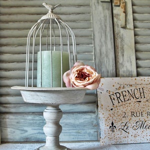 Könnte beinhalten: Ein weißer Metall-Kuchenständer mit einem Vogelkäfig und einer Kerze darauf. Eine rosa Rose liegt am Rand des Ständers. Der Ständer steht auf einer weißen Leinen-Tischdecke. Ein Schild mit dem Text "French 2. Rue Le Me" befindet sich im Hintergrund.