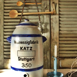 Może przedstawiać: Antyczny biały ceramiczny dzbanek z niebieskimi ozdobami i napisem "Weinessigfabrik KATZ Stuttgart 350" z przodu. Dzbanek ma dzióbek na dole i stoi na drewnianej powierzchni.