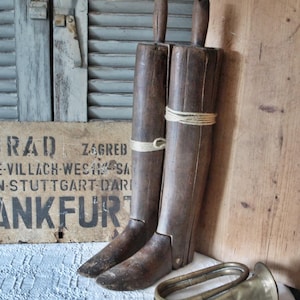 Puede incluir: Dos hormas de bota de madera antiguas atadas con cuerda, de pie. Una trompeta de latón vintage descansa sobre un mantel de encaje blanco. Un letrero de madera desgastado y una tabla de madera están en el fondo.