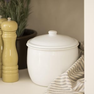 Op de afbeelding: Een witte emaille pot met deksel, die op een wit rek staat met een gestreepte doek. Twee gele pepermolens staan op het rek voor de pot.