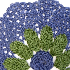 Könnte beinhalten: Eine gehäkelte Deckchen mit einer blauen Mitte und grünen Blättern am Rand. Die Deckchen ist mit einem zarten Stichmuster gearbeitet.
