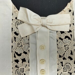Könnte beinhalten: Eine weiße Bluse mit Spitzenbesatz und einer Schleife aus Satin am Ausschnitt. Die Bluse hat drei Knöpfe vorne.