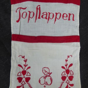 Könnte beinhalten: Weißes Stoffküchenhandtuch mit roter Stickerei, die "Topflappen" sagt, und einem roten Stickmuster mit zwei Pflaumen und zwei Blumen.