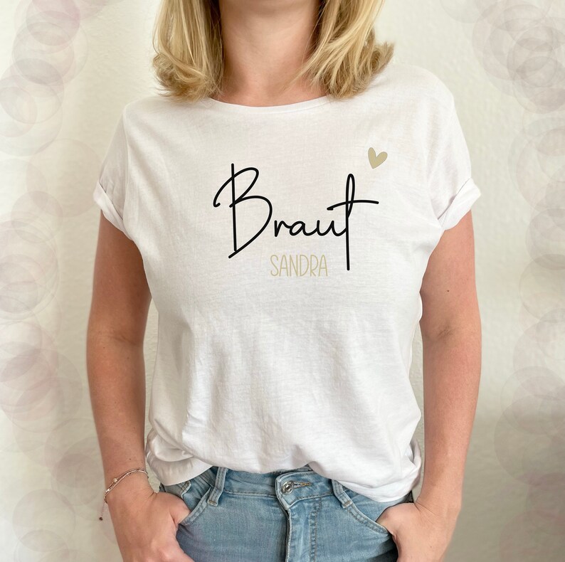 Pode incluir: Camiseta branca com texto preto que diz "Braut Sandra" com um pequeno cora&ccedil;&atilde;o dourado sobre o texto.