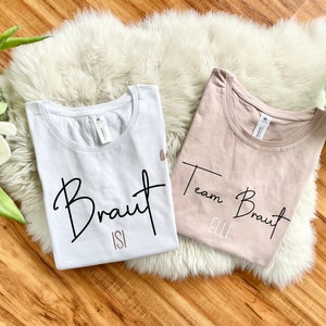 Pode incluir: Duas camisetas brancas com texto preto. Uma camiseta diz "Braut Isi" e a outra diz "Team Braut Elli".