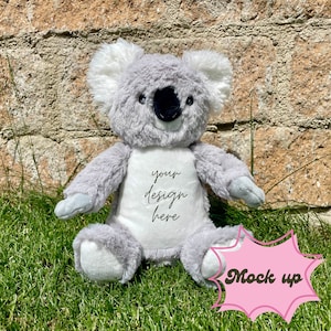 May include: A gray plush koala bear with white accents on its ears and paws, sitting on green grass. The koala has a black nose and the text "your design here" is embroidered on its chest. A pink "Mock up" graphic is in the bottom right corner.