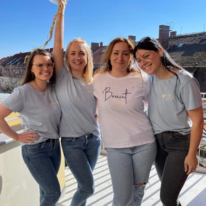 Pode incluir: Quatro mulheres vestindo camisetas azuis claras, uma delas com a palavra "Braut" na camiseta. Elas est&atilde;o segurando bal&otilde;es e sorrindo.