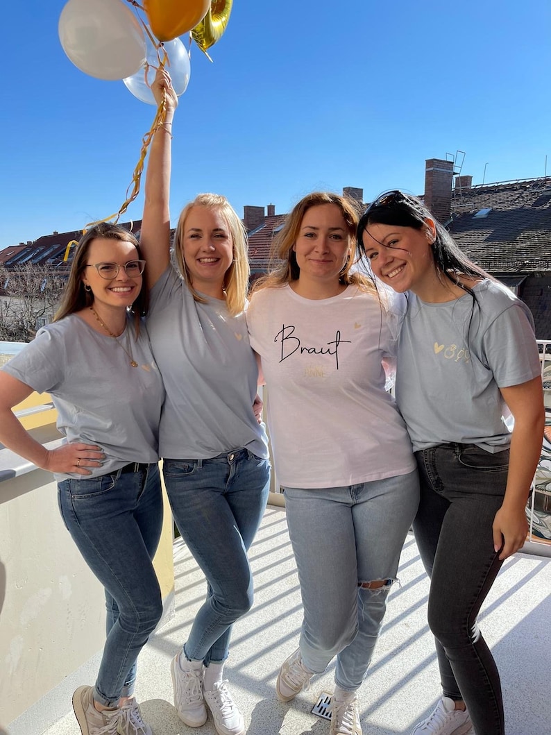 Pode incluir: Quatro mulheres vestindo camisetas azuis claras, uma delas com a palavra "Braut" na camiseta. Elas est&atilde;o segurando bal&otilde;es e sorrindo.