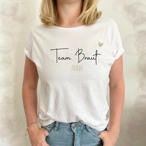 Pode incluir: Camiseta branca com texto preto que diz "Team Braut Anna" e um pequeno cora&ccedil;&atilde;o dourado.