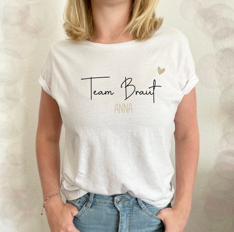 Pode incluir: Camiseta branca com texto preto que diz "Team Braut Anna" e um pequeno cora&ccedil;&atilde;o dourado.