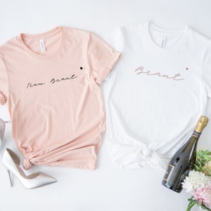 Pode incluir: Duas camisetas brancas com texto preto. Uma camiseta diz "Team Bride" e a outra diz "Bride". Ambas as camisetas t&ecirc;m um pequeno cora&ccedil;&atilde;o preto ao lado do texto.