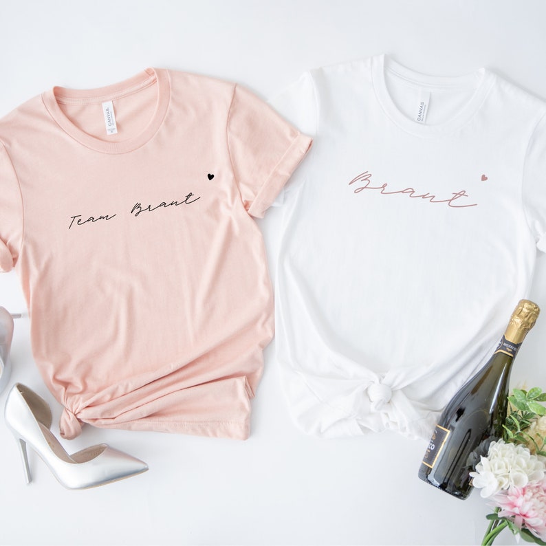 Pode incluir: Duas camisetas brancas com texto preto. Uma camiseta diz "Team Bride" e a outra diz "Bride". Ambas as camisetas t&ecirc;m um pequeno cora&ccedil;&atilde;o preto ao lado do texto.