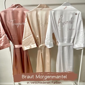 Könnte beinhalten: Drei Satin-Morgenmäntel in verschiedenen Farben, mit den Worten "Trauzeugin", "Mrs. Ray" und "Braut Desiree" auf der Rückseite bestickt. Die Mäntel hängen an einem weißen Kleiderständer.