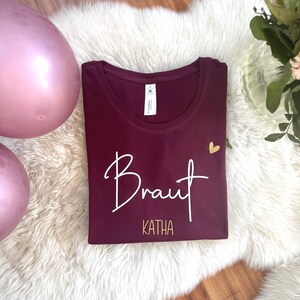 Pode incluir: Camiseta cor vinho com texto branco que diz "Braut Katha" e um pequeno cora&ccedil;&atilde;o dourado.