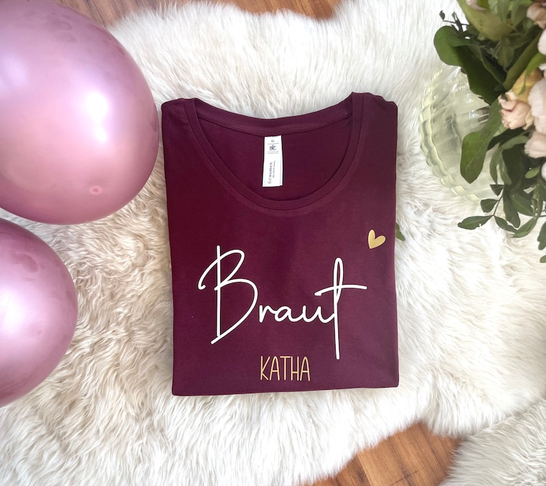 Pode incluir: Camiseta cor vinho com texto branco que diz "Braut Katha" e um pequeno cora&ccedil;&atilde;o dourado.