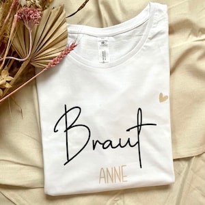 Pode incluir: Camiseta branca com a palavra "Braut" em escrita preta e "ANNE" em escrita dourada abaixo. Um pequeno cora&ccedil;&atilde;o dourado est&aacute; no canto superior direito.