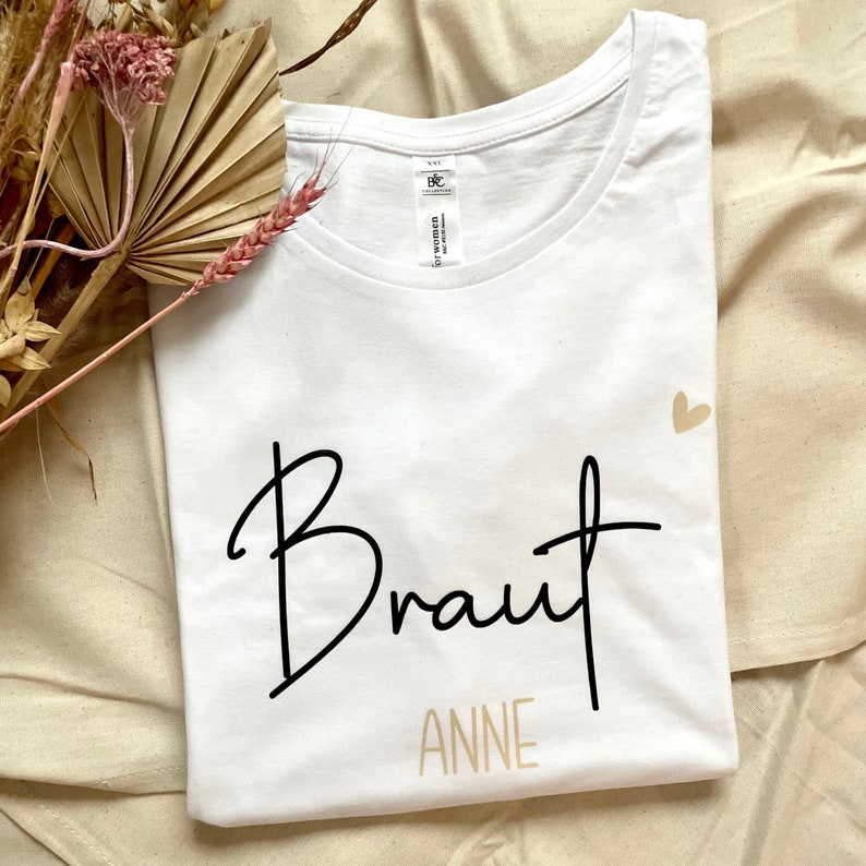 Pode incluir: Camiseta branca com a palavra "Braut" em escrita preta e "ANNE" em escrita dourada abaixo. Um pequeno cora&ccedil;&atilde;o dourado est&aacute; no canto superior direito.