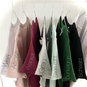 Pode incluir: Oito t-shirts penduradas em cabides, mostrando diferentes cores: branco, rosa claro, bord&ocirc;, cinza, verde claro, verde escuro, preto. As t-shirts s&atilde;o etiquetadas com as seguintes cores: Wei&szlig;, Altros&eacute;, Weinrot, Grau, Sage-mint, Dunkelgr&uuml;n, Schwarz.